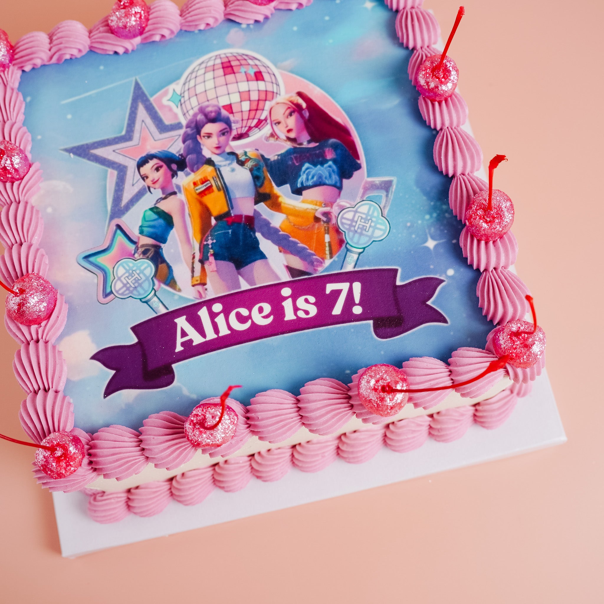 Birthday cake with pink decorations and a picture of huntrix, the band from k-pop demon hunters on a blue background, celebrating &