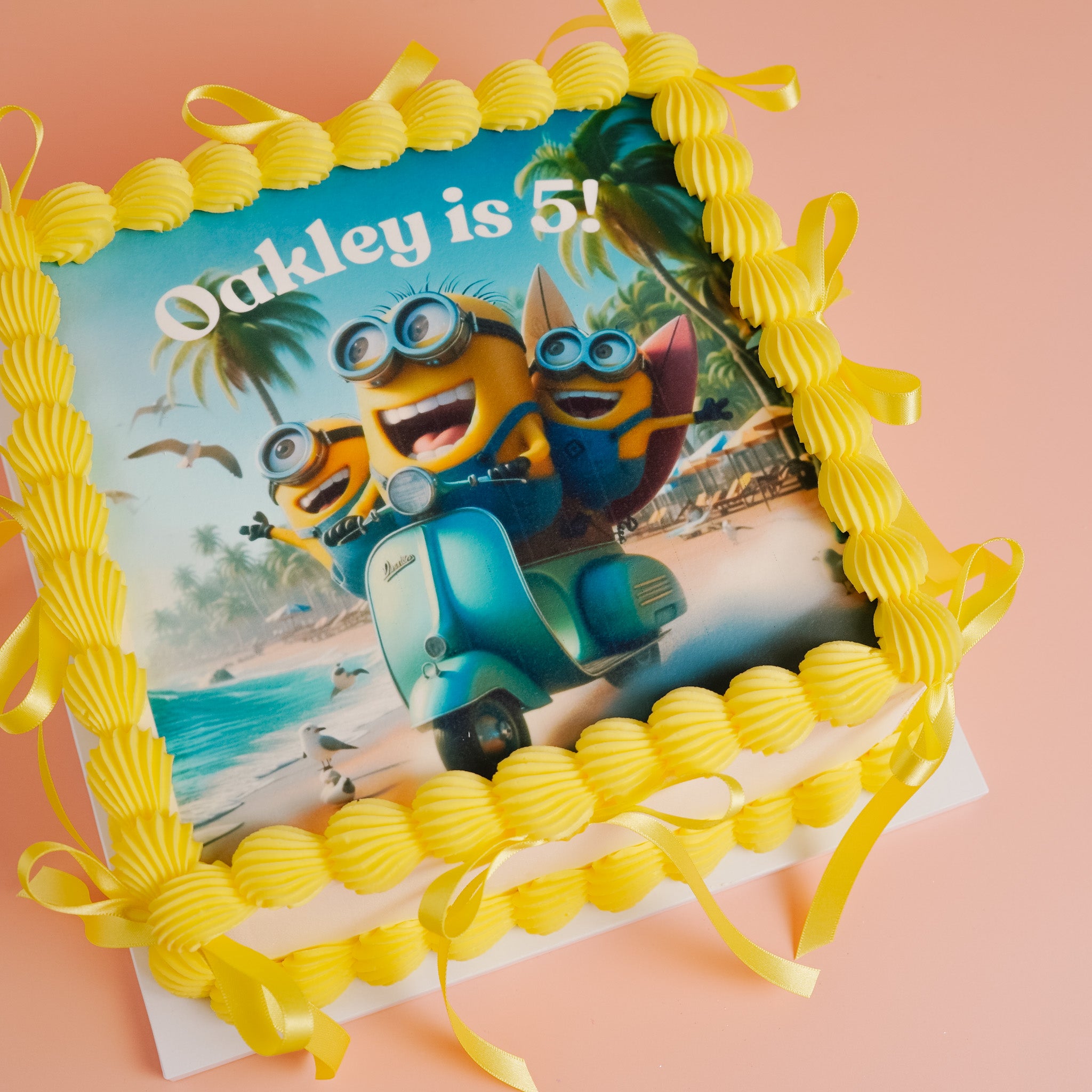 Birthday cake with Minions cake topper on a scooter, on a square vanilla cake, personalised for Oakley, photographed on a pink background.