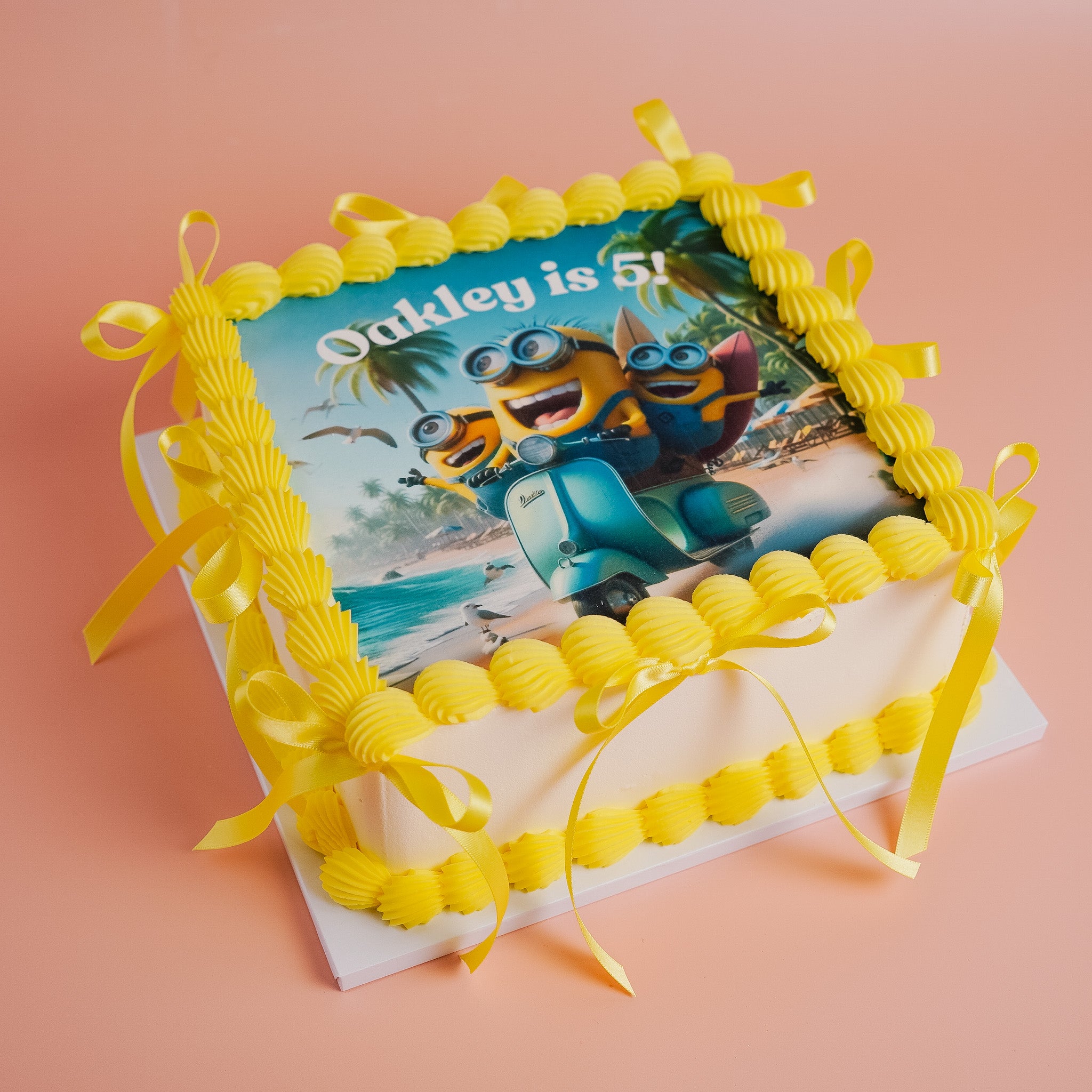 Vanilla sqaure cake with Minions design and yellow buttercream shell and yellow ribbon on a pink background