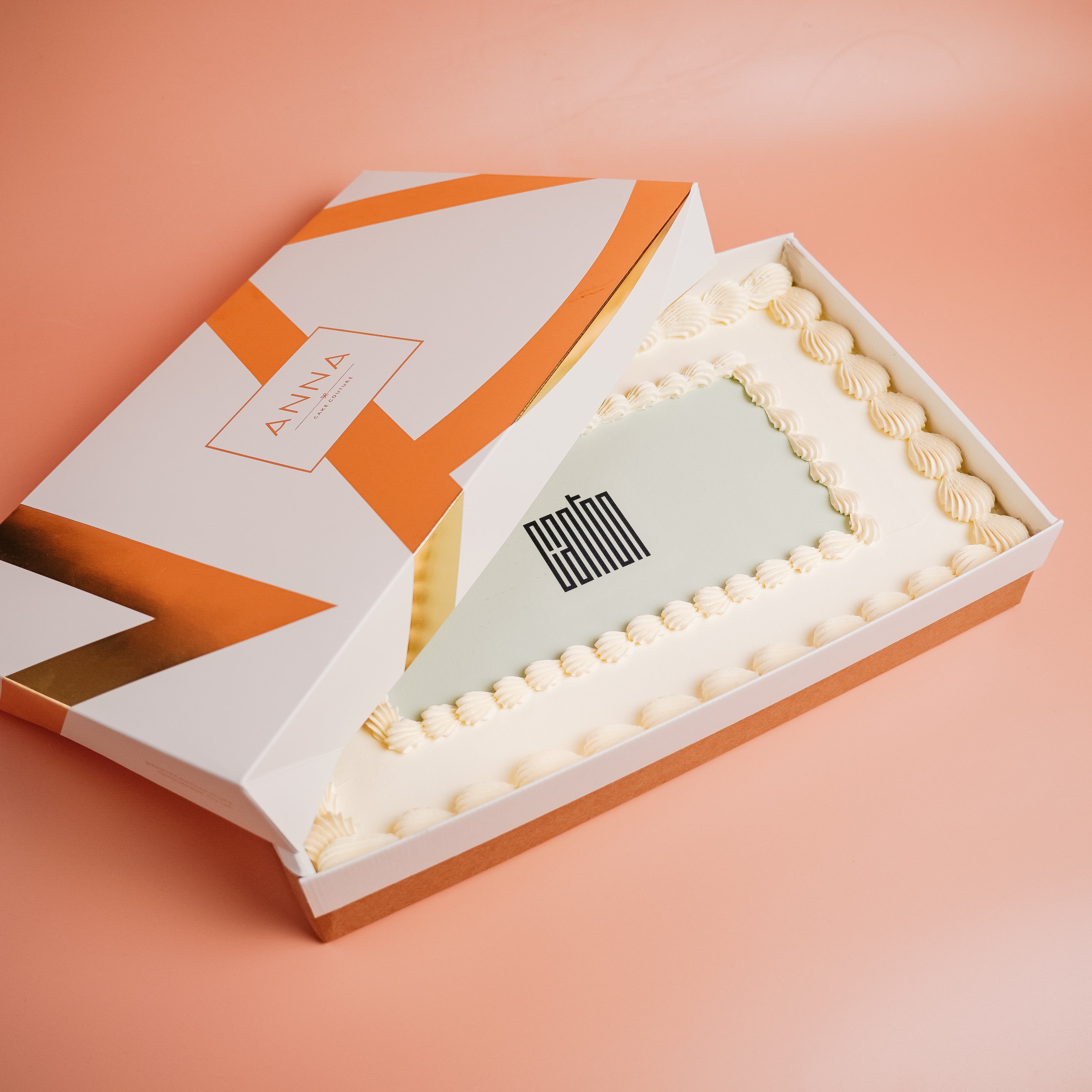 Open box containing a cake with decorative elements and a printed logo on a peach background