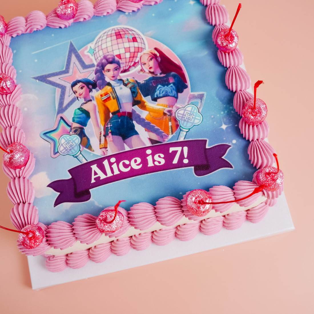 Birthday cake with pink decorations and a picture of huntrix, the band from k-pop demon hunters on a blue background, celebrating &