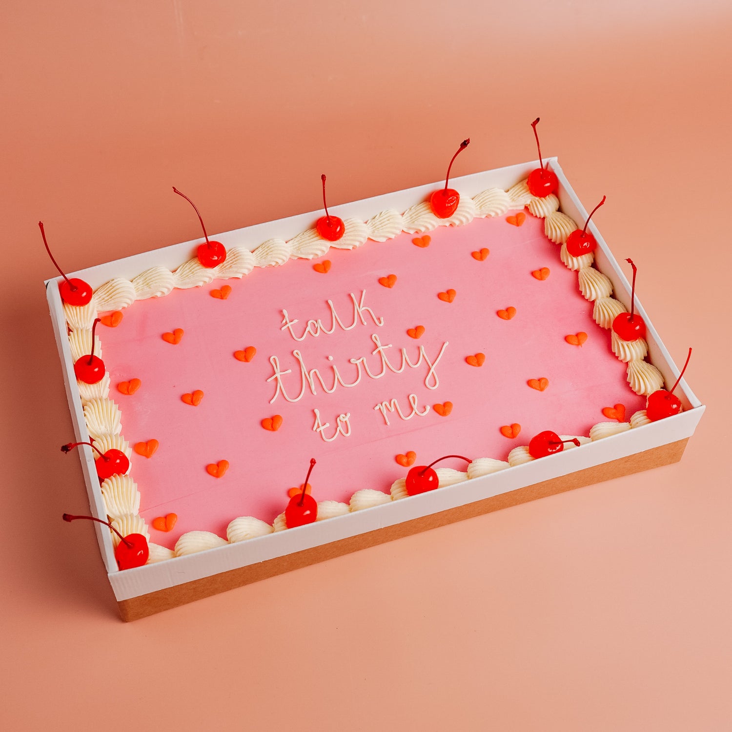 Decorative cake with pink icing, white accents, red piped hearts and red cherries on a peach background.